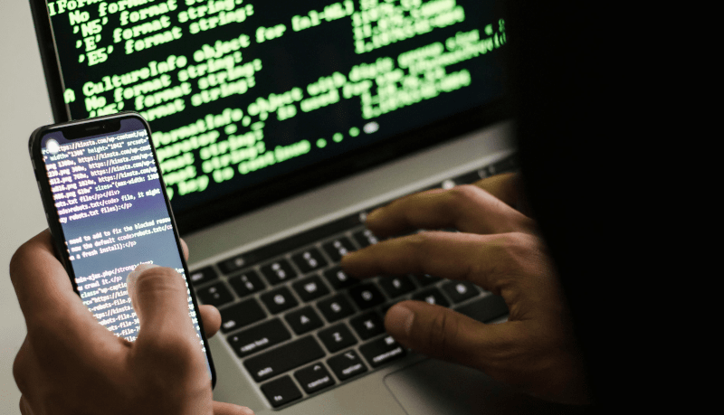 Imagem de um hacker digitando em um laptop com a  mão direita e segurando um smartphone na mão esquerda.