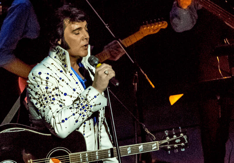 Elvis cantando em pé de olhos fechados, com a guitarra pendendo do pescoço